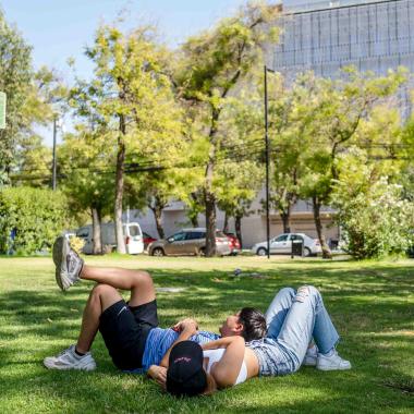 Dos estudiantes acostadas en el pasto dentro del campus universitario. Al lado se situa un letrero que indica zona de descanso.