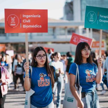 Dos estudiantes utilizando poleras que indican "Estudia en la Usach". Una lleva un cartel de Ingeniería Civil Biomédica en la mano y la otra estudiante lleva un cartel de Terapia Ocupacional.