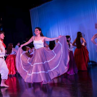 Sobre el escenario una pareja de estudiantes bailan sonrientes música folclórica venezolana. Detrás hay un grupo de mujeres que acompañan bailando.