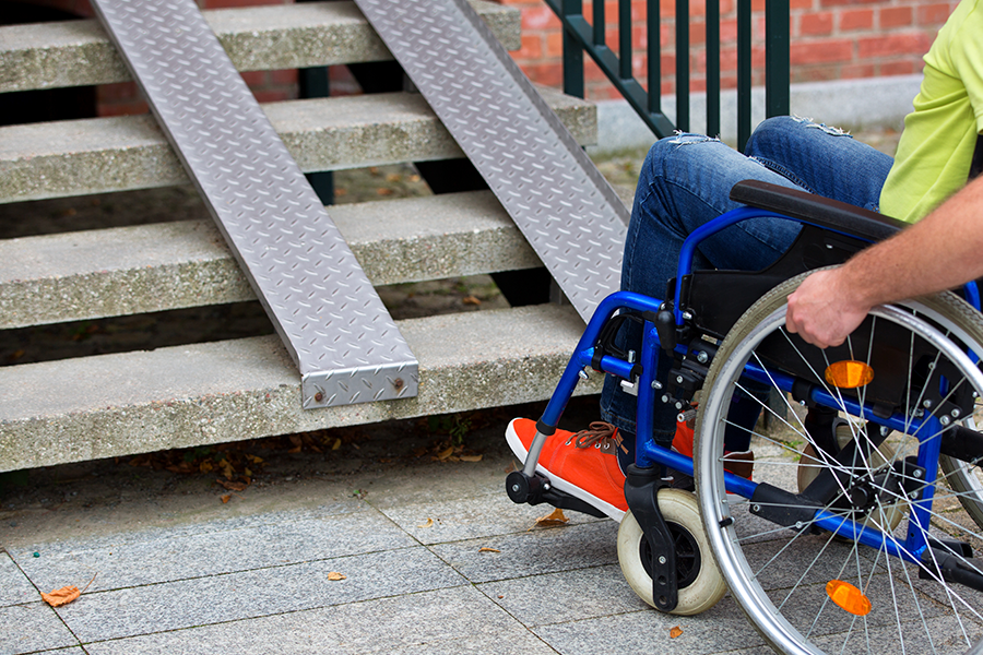A person in a wheelchair is assisted on a sidewalk next to a concrete staircase with a portable metal ramp extending over a few steps.