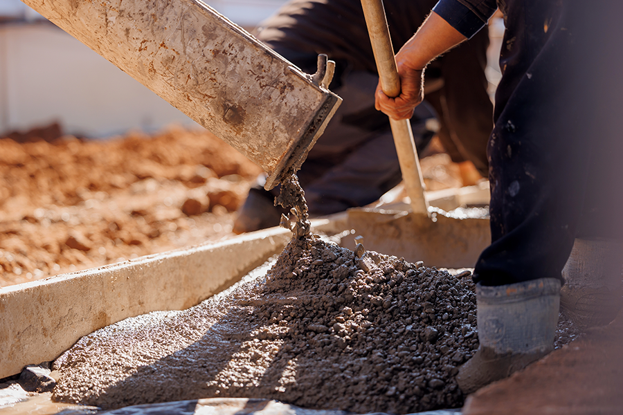 A close-up captures construction workers pouring wet, grainy concrete from a bucket onto a flat base, with dry soil visible in the background.