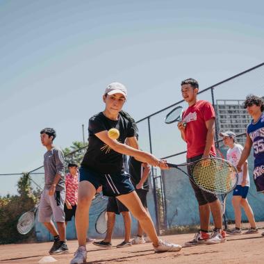 Plano general de mujer sosteniendo una raqueta de tenis a punto de pegarle a una pelota en el aire en una cancha. Detrás de ella hay siete personas más con raquetas de tenis en las manos.