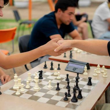Plano medio de dos jóvenes dándose la mano antes de comenzar una partida de ajedrez. Debajo de las manos se posiciona el tablero y en el costado se encuentra un cronómetro. 
