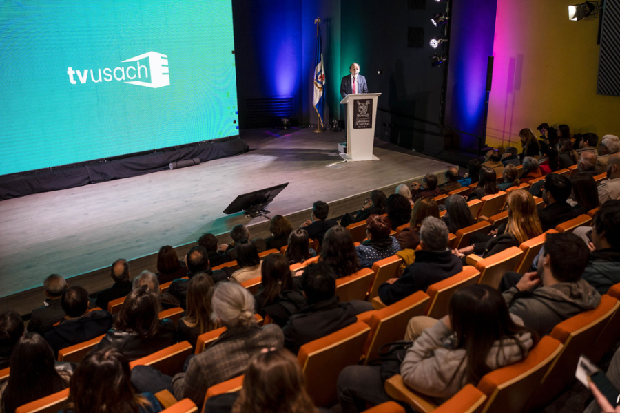 La fotografía exhibe el auditorio FAE lleno, una pantalla gigante con el nuevo logotipo de TV Usach y el Rector Vidal en el podio dirigiéndose a la audiencia.