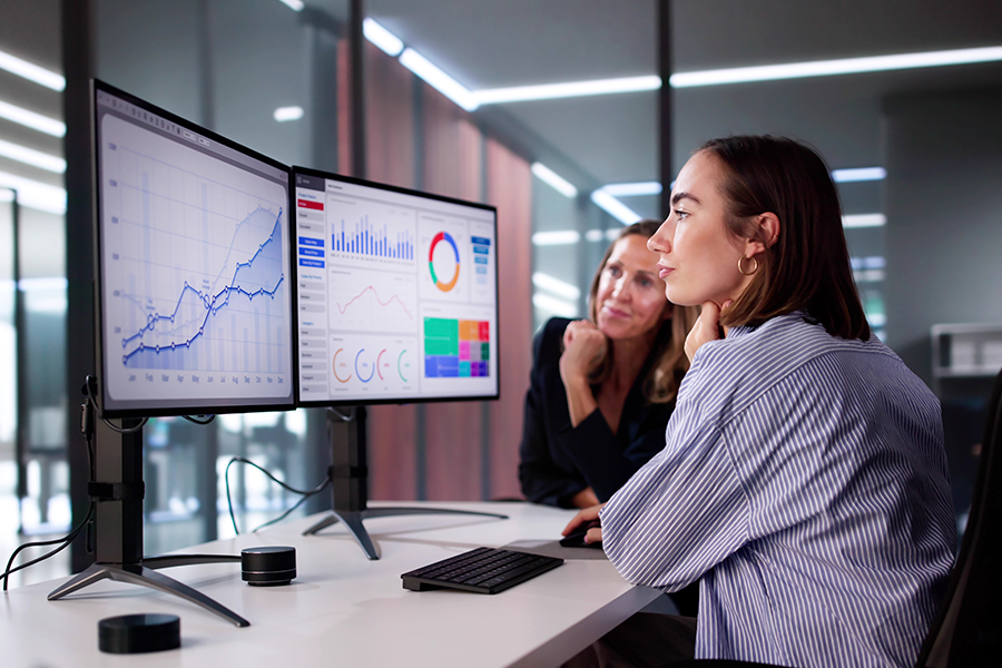 In a modern office, two professional women analyze market research on two monitors displaying data visualizations, tables, and graphs.