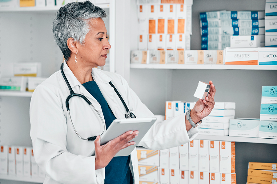 A doctor in a white coat and stethoscope holds a tablet and examines a box of medicines in front of shelves full of pharmaceutical products.
