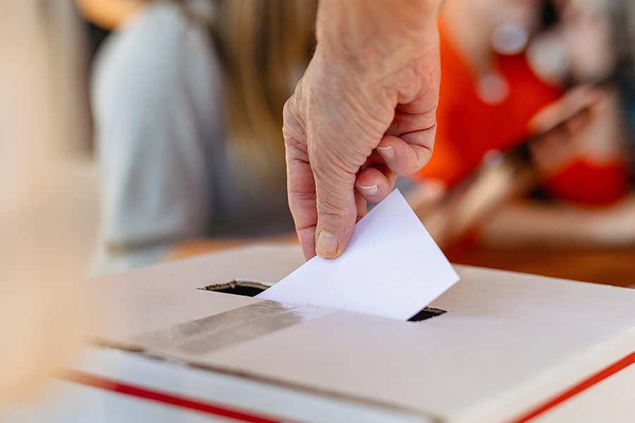 A hand, in the foreground, inserts a folded piece of paper through the slot of a ballot box.