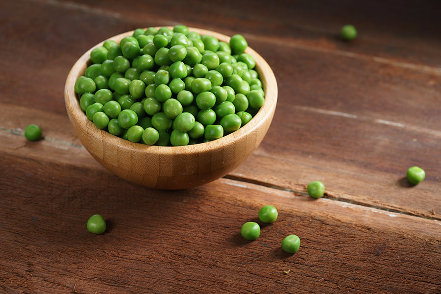 Un pequeño plato redondo de madera lleno de arvejas de color verde brillante se ubica sobre una superficie de madera, con varias arvejas esparcidas alrededor.