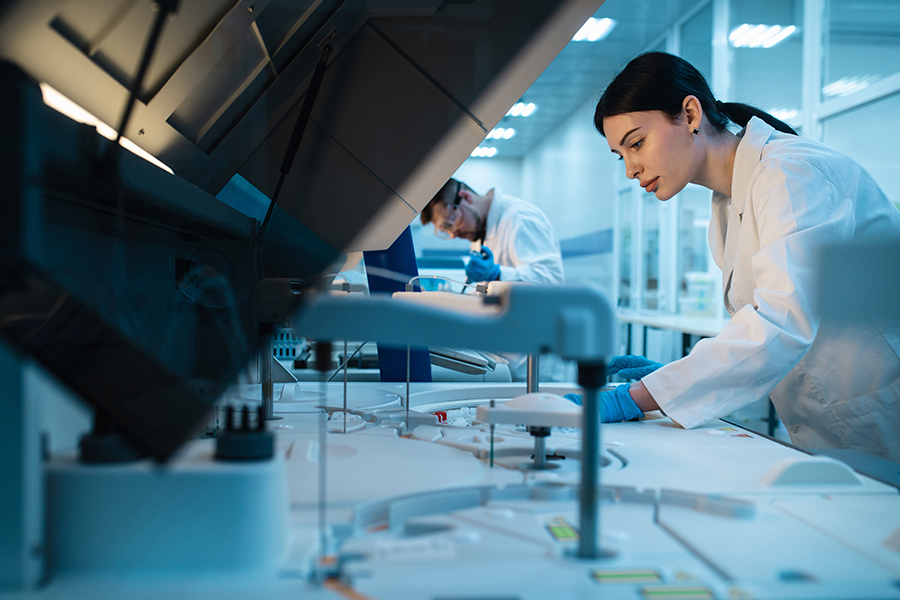 Two scientists wearing white coats and blue gloves work with advanced machinery in a modern, well-lit laboratory.