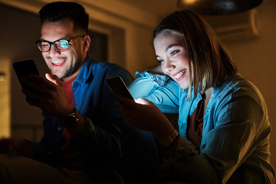 Una pareja sonriente se sienta muy junta en una habitación oscura, iluminada sólo por la luz de las pantallas de los smartphones que están mirando.