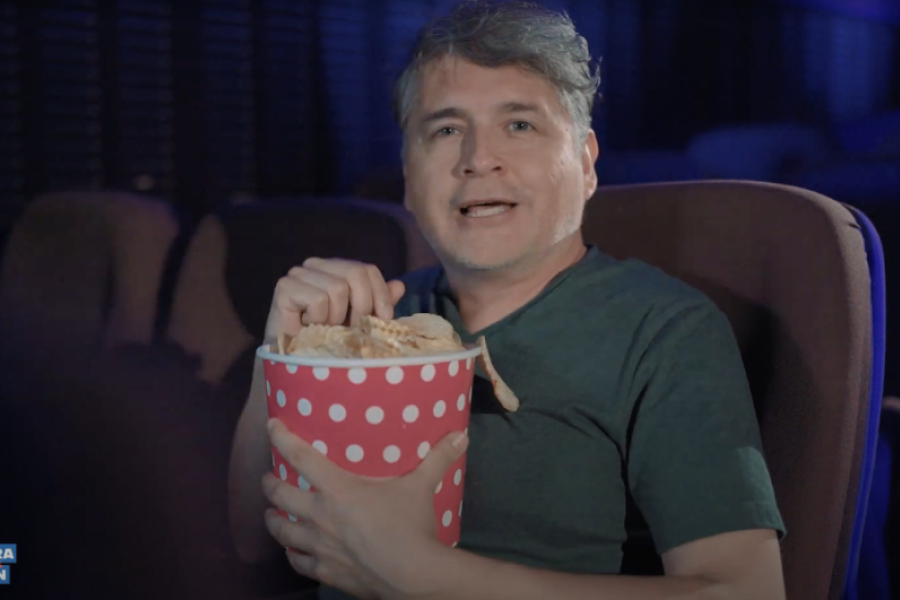 Hombre sentado en butacas mirando a la cámara con un paquete de palomitas en la mano
