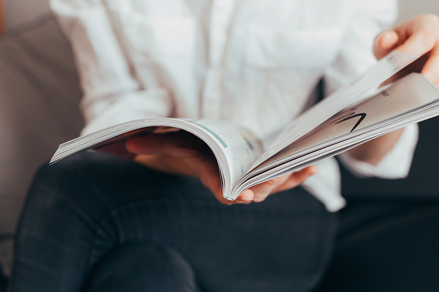 A person in a white shirt and dark pants sits and flips through the pages of a magazine, their hands holding the spine and turning a page.
