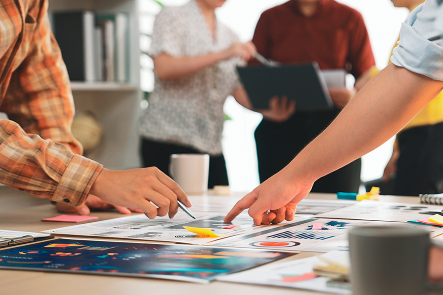 A close-up of a wooden table where several people are pointing at colorful data charts and world maps during a meeting.