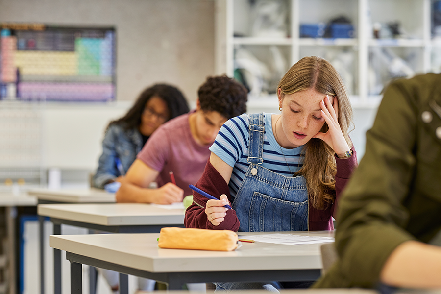 A student in a blue striped shirt and denim overalls focuses on an exam in a classroom while other students work in the background.