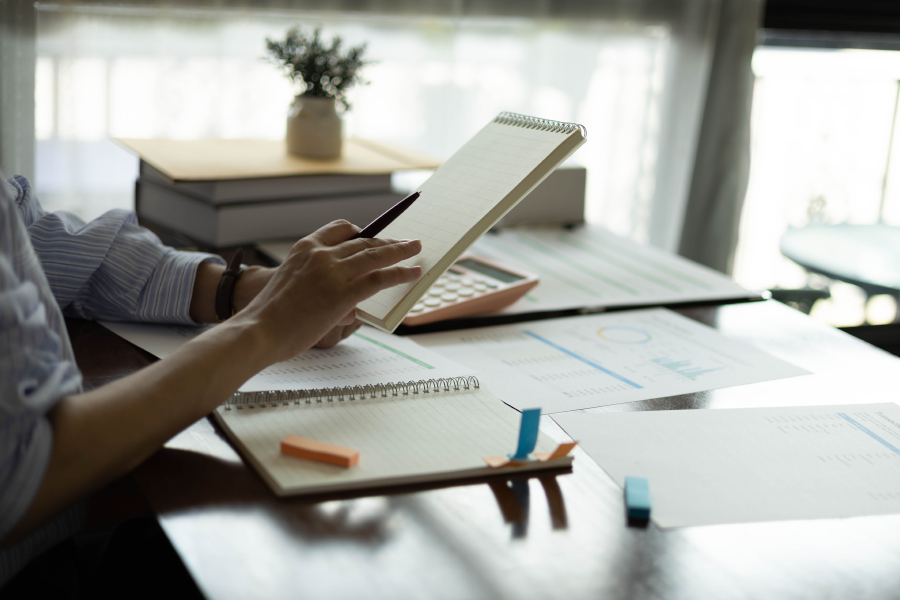 Person at a desk using a pen and calculator over various printed graphs and open notebooks.