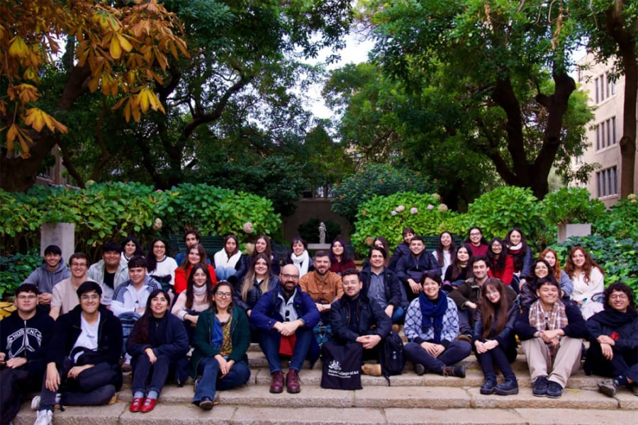 En la imagen aparecen los participantes del evento sentados sobre unas gradas del edificio en donde se celebró la actividad. De fondo se logra divisar áreas verdes.