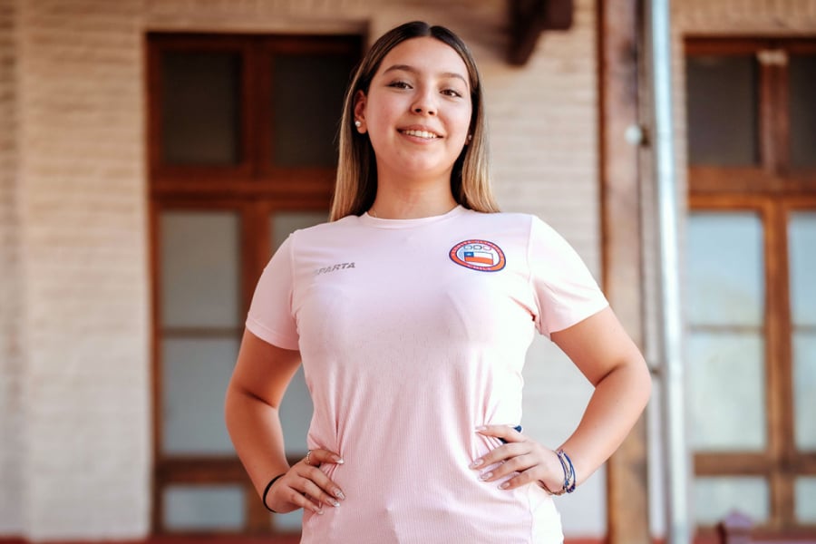 En l imagen aparece nuestra estudiantes seleccionada nacional de patinaje. Posa a la cámara con una polera blanca con el escudo del país en su costado izquierdo. De fondo la EAO. 