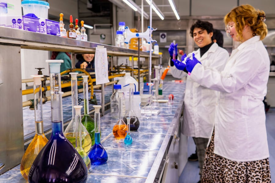 LA imagen muestra a dos estudiantes con cotona blanca y guantes azules trabajando en un laboratorio