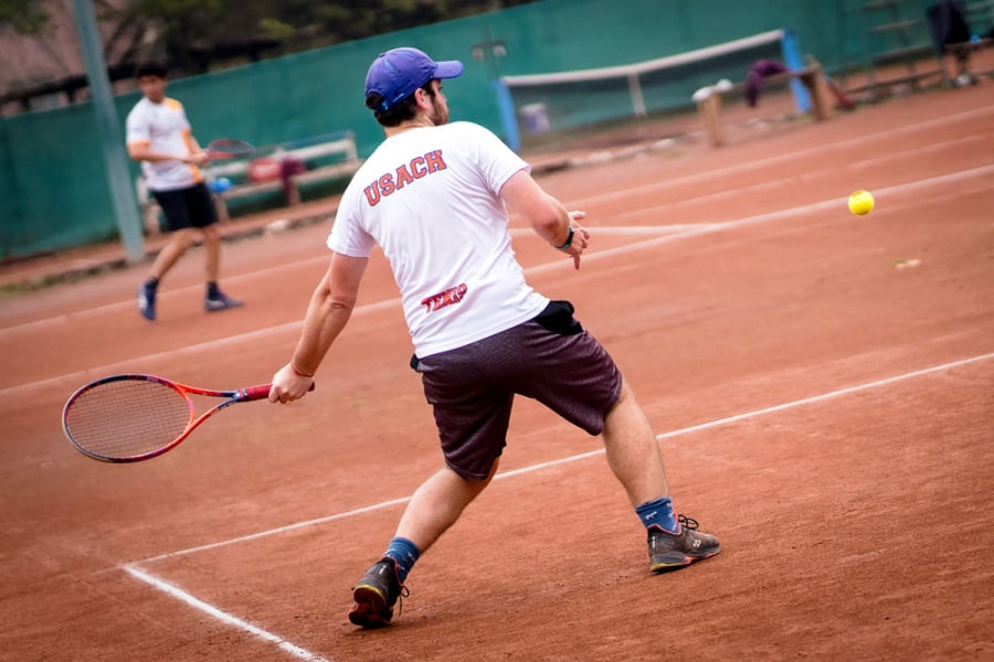 En la imagen se muestra a un estudiante de espaldas jugando tenis, con la raqueta en la mano izquierda, en una cancha de arcilla