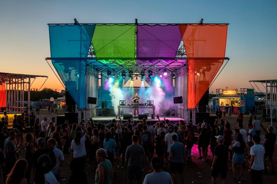 La foto muestra uno de los escenarios del evento. Con telas que adornan el espacio de colores celeste, verde, morado y naranjo y un hombre ubicado al centro del escenario con una mesa de sonido