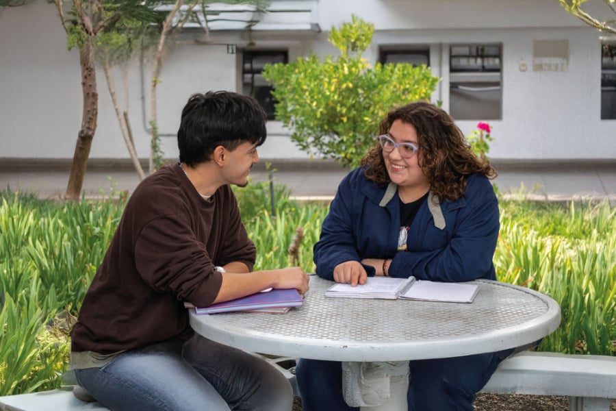 La foto muestra a dos estudiantes, un hombre y una mujer, sentados conversando en el patio de la facultad tecnológica