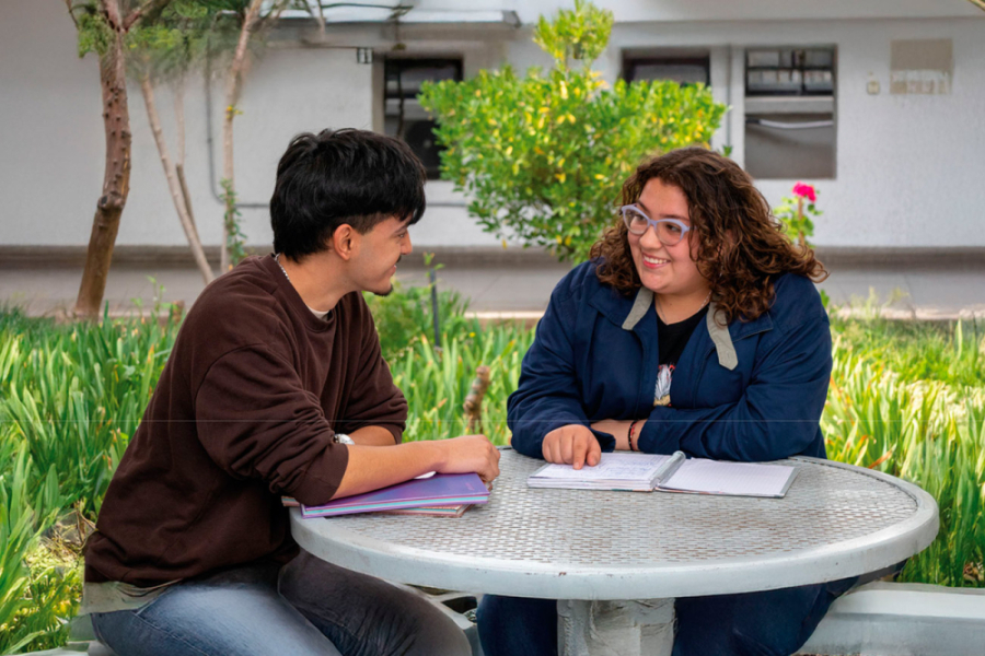 La foto muestra a dos estudiantes, un hombre y una mujer, sentados conversando en el patio de la facultad tecnológica