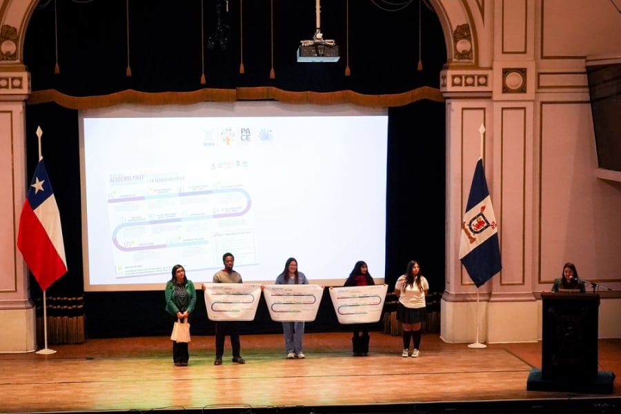 La imagen muestra a cinco personas, cuatro mujeres y un hombre. Tres de ellas sostienen una pancarta en el escenario del Aula Magna de la Usach