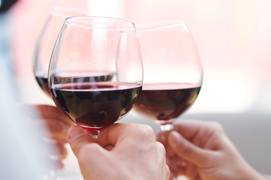 Manos brindando con tres copas de vino tinto sobre un fondo claro y difuminado.