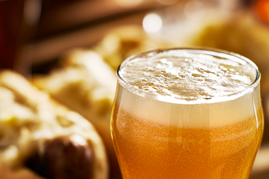 Vaso de cerveza rubia con abundante espuma en primer plano y comida difuminada atrás.