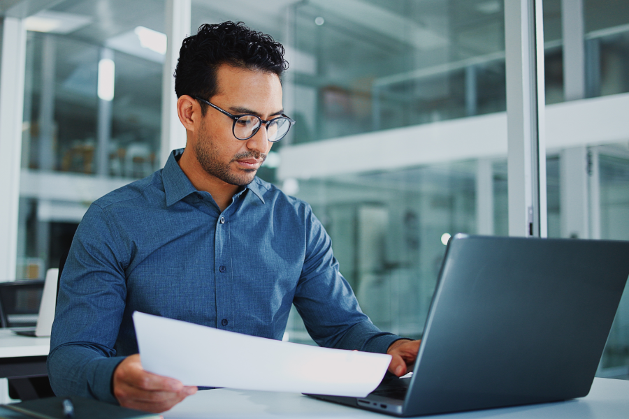 Un hombre con anteojos revisa un documento mientras trabaja en un computador laptop.
