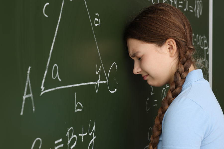 Niña con expresión de frustración apoya su frente contra una pizarra con un problema de geometría.