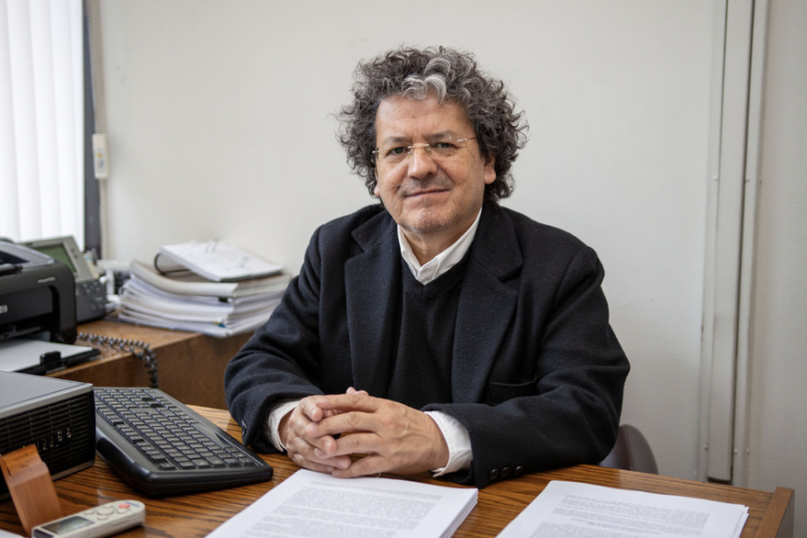 La foto muestra al académico sentado en su escritorio con las manos cruzadas sobre la mesa
