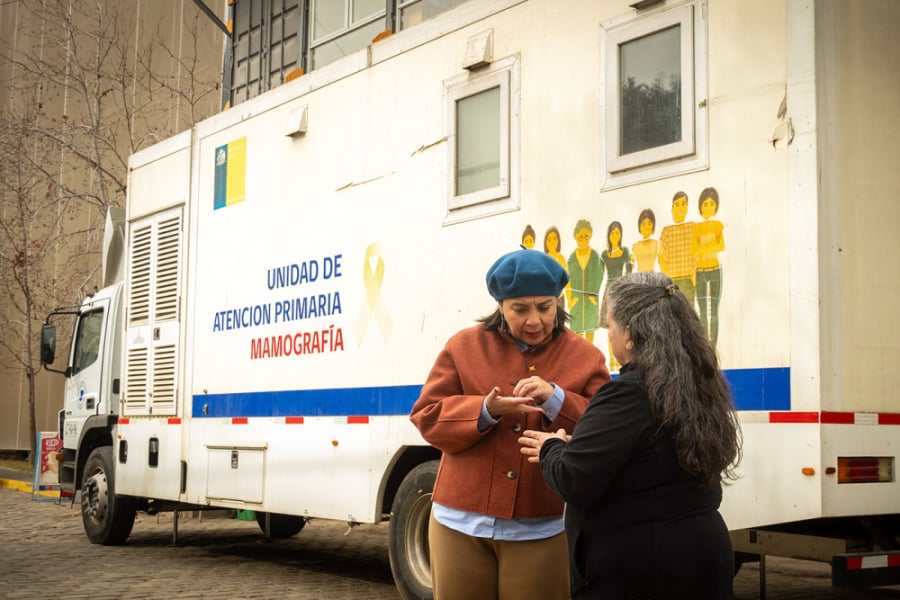 La foto muestra a dos mujeres conversando frente al vehículo de atención primaria de mamografía 