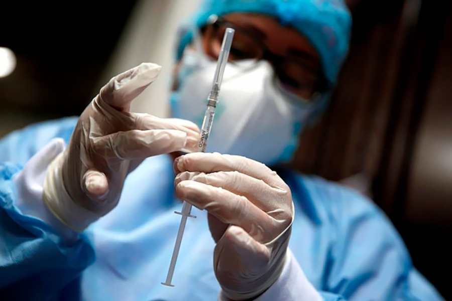 La imagen muestra a un médico sosteniendo una jeringa. Con guantes quirúrgicos, cofia, mascarilla y bata celeste 