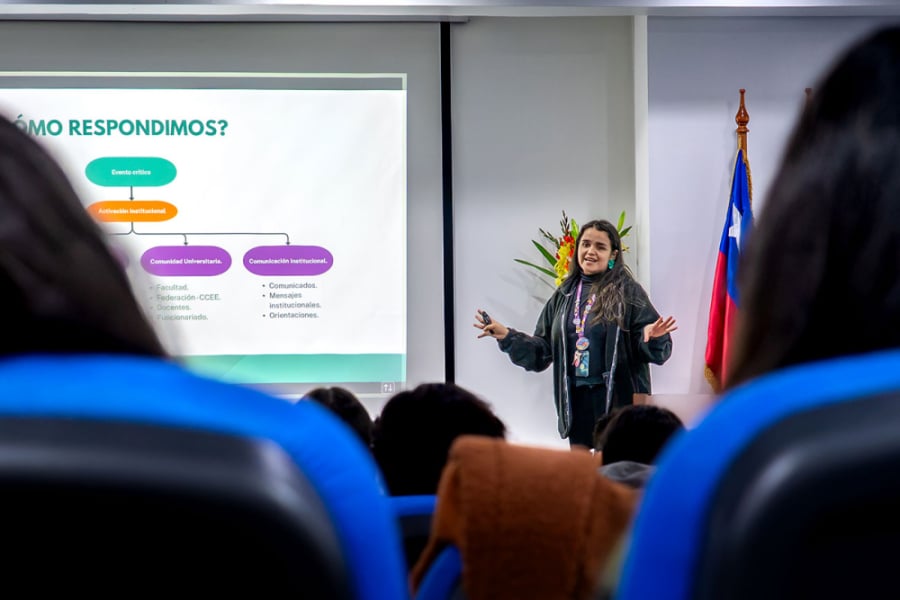 La imagen muestra a una de las participantes de la dirección, dictando una charla. Con sus brazos abiertos y de fondo la presentación del proyecto